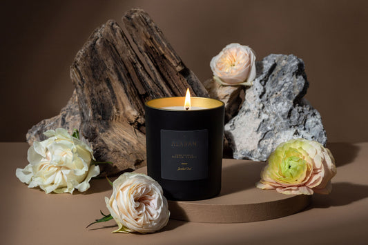 Jardin Oud lit candle next to light pink and white roses on a brown background